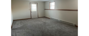 an empty living room with gray carpet and two windows and an entry door