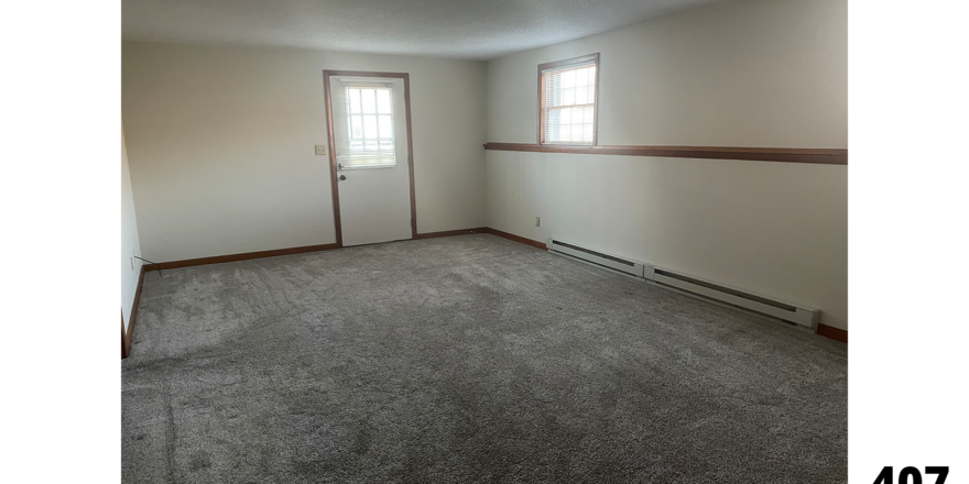 an empty living room with gray carpet and two windows and an entry door